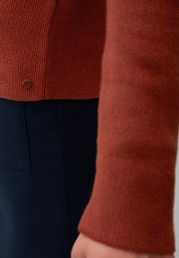 Strickpullover in Rostfarbe mit einem strukturierten Muster, der einen abgerundeten Saum und einen Metalllogoschmuck am unteren Rand aufweist. Kombiniert mit einem dunklen Rock.