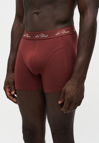 Buste masculin et haut des jambes portant des boxers ajustés rouge foncé avec une ceinture marquée affichant "Les Deux" sur un fond clair uni.