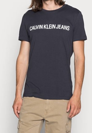Person trägt ein dunkelblaues T-Shirt mit weißem Aufdruck „CALVIN KLEIN JEANS“ auf der Brust und beige Cargohosen.