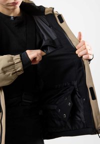 Die Innenausstattung der tan- und schwarzen Jacke verfügt über mehrere Taschen, ein glattes Futter und verstellbare Riemen. Der Außenstoff wirkt strukturiert und langlebig.