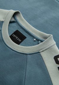 Sudadera azul con cuello redondo, que presenta un acento en azul claro y verde. La tela parece suave con una etiqueta que dice "ombre".