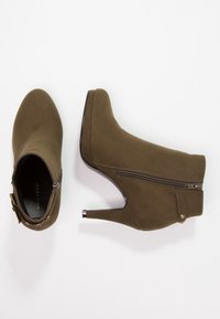 Bottines en daim vert olive avec un talon aiguille, une fermeture éclair sur le côté et des accents en métal décoratifs. Texture lisse et design à bout pointu.
