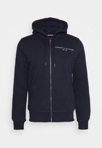 Sudadera con cremallera en color navy, hecha de algodón suave. Cuenta con un bolsillo frontal, capucha con cordón y un logotipo prominente en el pecho. Textura suave.