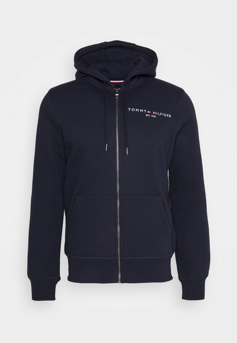 Sudadera con cremallera en color navy, hecha de algodón suave. Cuenta con un bolsillo frontal, capucha con cordón y un logotipo prominente en el pecho. Textura suave.