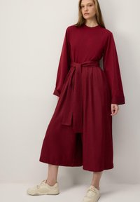 Bordové šaty s dlhým rukávom a opaskom v páse, s voľným spodkom v štýle culottes a jemnou textúrou. Kombinované s ľahkými teniskami.