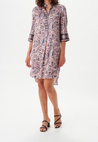 Kobieta w sukience do kolan z długim rękawem, ozdobionej różowo-fioletowym wzorem paisley, oraz w czarnych sandałach na obcasie, stojąca na białym tle.