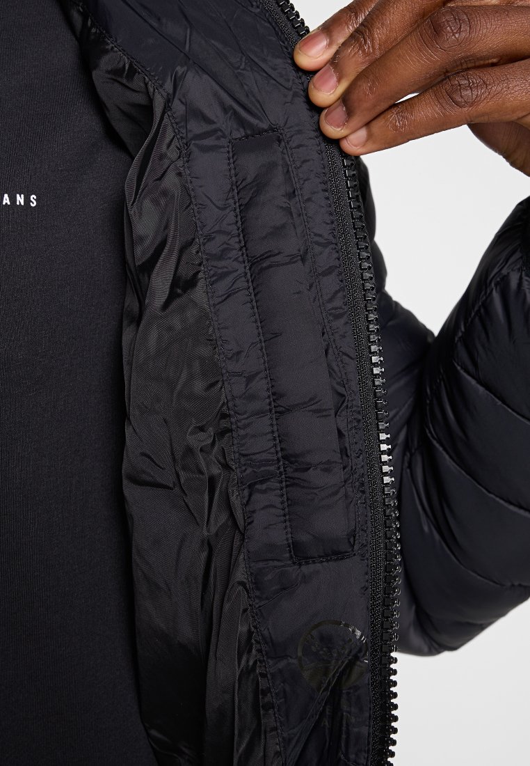 Doudoune noire avec une texture lisse et brillante, dotée d'une fermeture éclair et d'une poche intérieure. Le tissu semble léger et isolant.