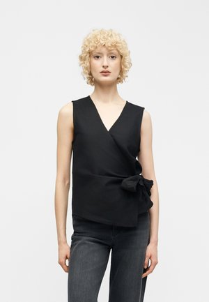 Ξανθιά γυναίκα με σγουρά μαλλιά, φοράει μαύρο αμάνικο τοπ με δέσιμο στο πλάι και σκούρο γκρι τζιν, στέκεται μπροστά σε απλό φόντο.