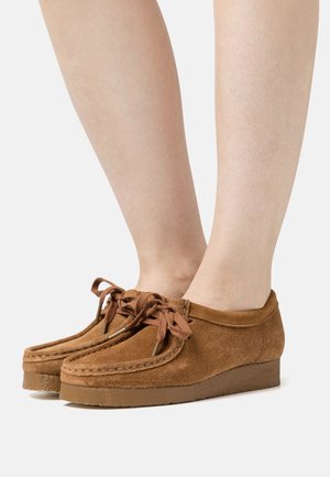 Chaussures mocassins à lacets en daim marron portées sur une paire de jambes à peau claire sur un fond uni.