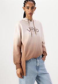 Bež i smeđi ombre hoodie s logotipom, kapuljača sa špagama i bočnim otvorima. Upareno s svijetloplavim trapericama. Mekani tkaninski motiv.