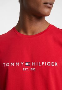 Camiseta de algodón roja con el texto bordado en blanco "TOMMY HILFIGER EST. 1985" y un pequeño acento de logotipo en azul, blanco y rojo. Cuello redondo.