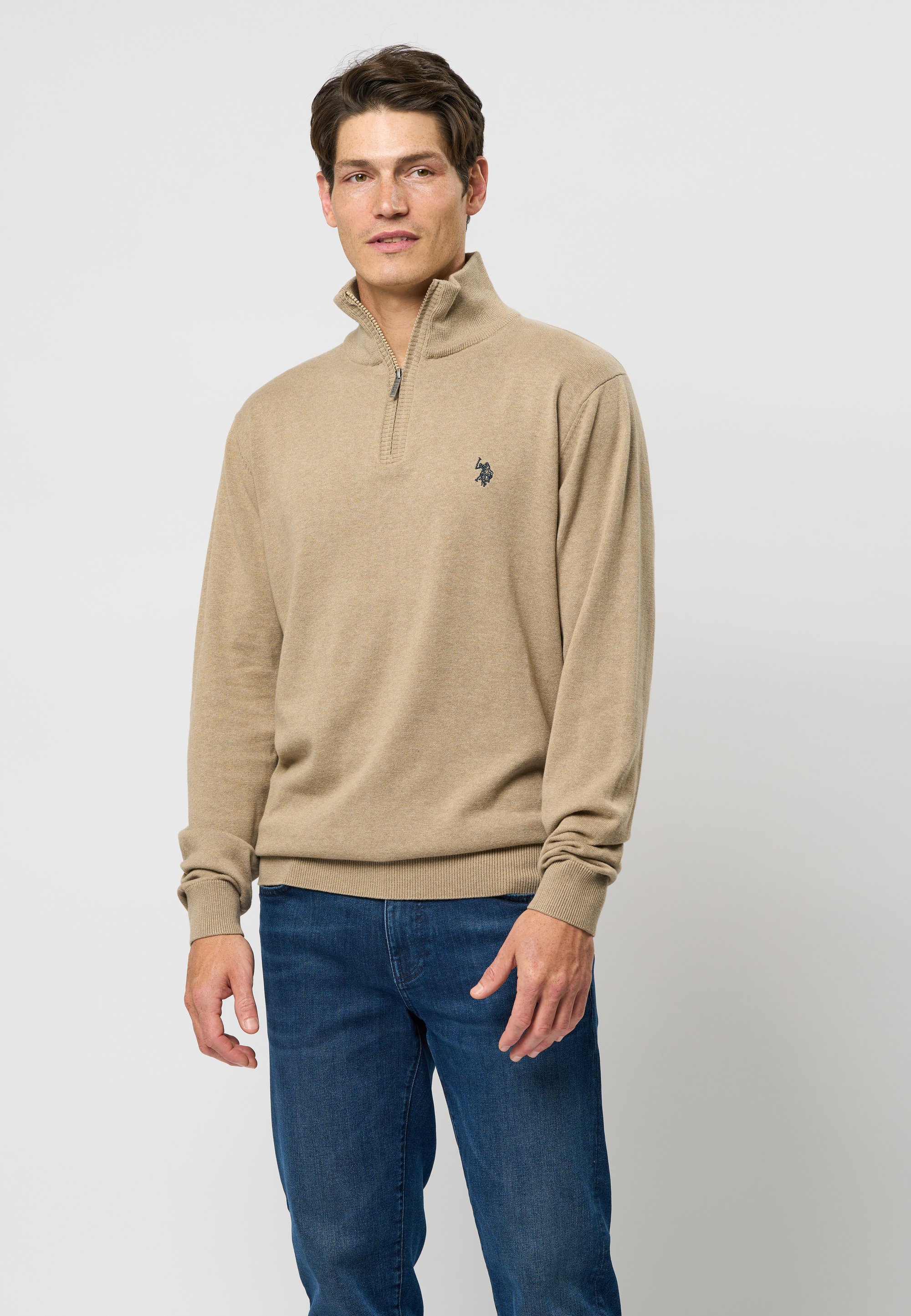 Man wearing tan quarter-zip sweater and blue jeans standing against plain light background.