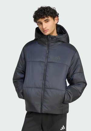 Junger Mann mit lockigem Haar, der eine schwarze Kapuzen-Steppjacke und schwarze Adidas-Hose trägt, mit den Händen in den Taschen vor einem schlichten Hintergrund.