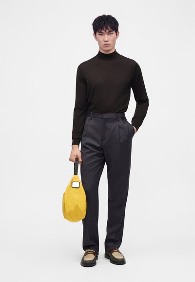 Homme portant un col roulé noir et un pantalon sombre tenant un sac jaune, debout devant un fond clair uni.