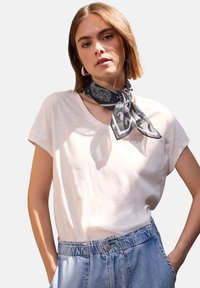 Hvid, løs t-shirt lavet af letvægtsstof, kombineret med et sort mønstrede tørklæde bundet om halsen og denimbukser med elastisk talje.