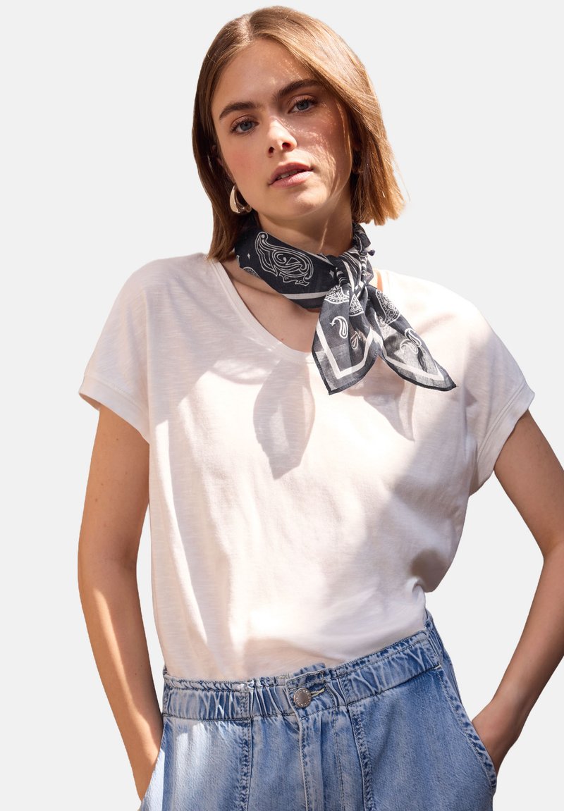 Hvid, løs t-shirt lavet af letvægtsstof, kombineret med et sort mønstrede tørklæde bundet om halsen og denimbukser med elastisk talje.