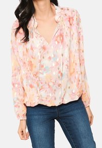 Kvinna i en pastellfärgad blus med blommönster och guldmetallic prickar, volang vid halsen och långa ärmar, kombinerad med mörkblå jeans.