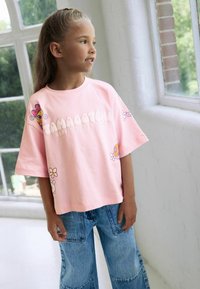Lyserød bomulds T-shirt med oversized pasform, med blomstergraphics og "Tamagotchi" tekst. Sættes sammen med lyseblå denim jeans.
