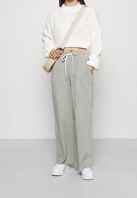 Gekürzter, weißer gestrickter Pullover mit Zopfmuster, kombiniert mit hellgrünen, weit geschnittenen Hosen mit Kordelzug und Seitentaschen. Weiße Sneakers.