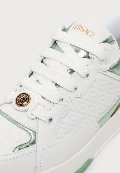 Λευκό αθλητικό παπούτσι Versace με χοντρές λευκές κορδόνια, χρυσό έμβλημα Μέδουσα, ανάγλυφο σχέδιο στο πλάι και μεταλλικές πράσινες λεπτομέρειες.