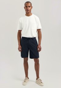 Tricou alb cu mânecă scurtă și buzunar, purtat cu pantaloni scurți bleumarin cu talie elastică, completat cu adidași bej.