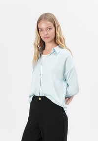 Lichtblauw overhemd met knopen in een zachte stof, met een kraag en een ontspannen pasvorm, gecombineerd met zwarte hooggesneden broek. Gouden knoopdetails.