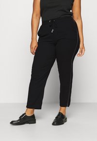 Zwarte joggers van zacht materiaal, met een tailleband met trekkoord en een witte zijkant. Er zijn zakken op heuphoogte.