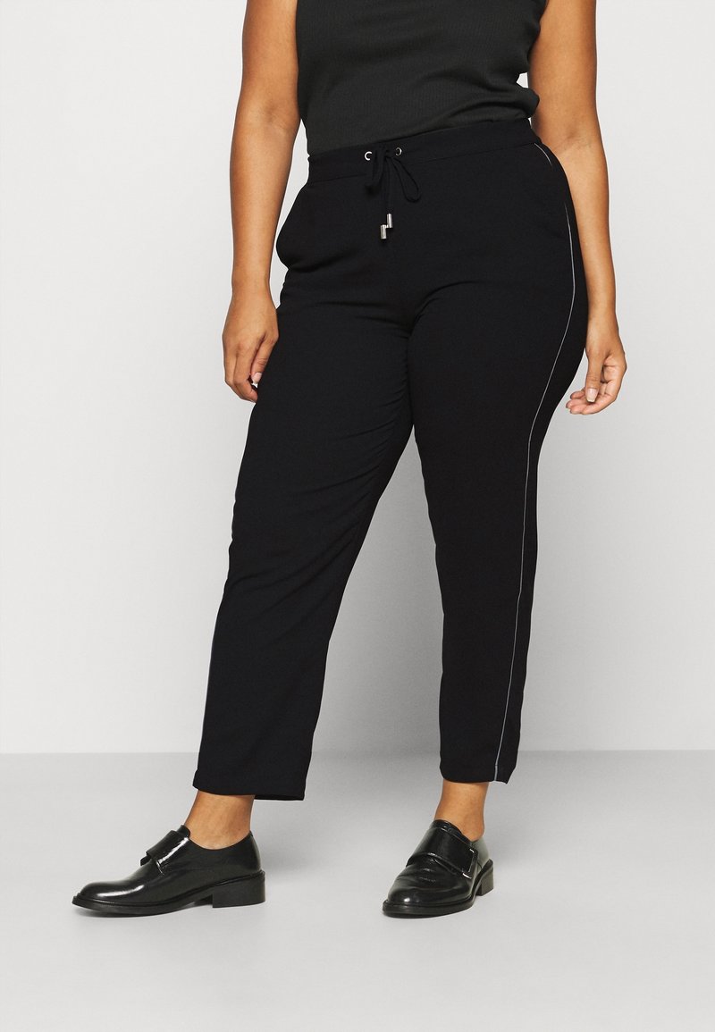 Zwarte joggers van zacht materiaal, met een tailleband met trekkoord en een witte zijkant. Er zijn zakken op heuphoogte.