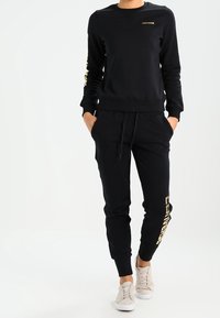 Persona che indossa un felpa nera con logo dorato e pantaloni jogger abbinati, con scarpe bianche, mani nelle tasche, in piedi su uno sfondo bianco.