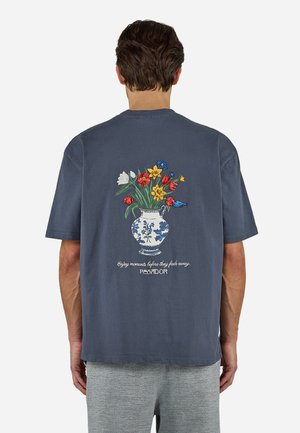 Άνδρας που φοράει σκουρόγριφο μπλουζάκι με πολύχρωμο γραφικό με βάζο με λουλούδια και λευκό κείμενο στην πίσω πλευρά, συνδυασμένο με γκρι παντελόνι με σχέδια.