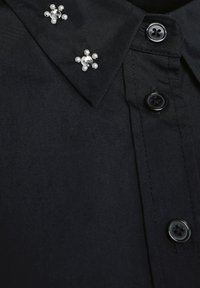 Camicia nera con collo appuntito decorato con due gruppi decorativi di perle finte e strass, e bottoni neri lungo la patta.