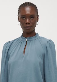 Lichtblauwe blouse met een hoge halslijn, gerimpelde kraag en sleutelgatopening. Bubbelsleeves voegen volume toe, terwijl de gladde stof een subtiele textuur heeft.
