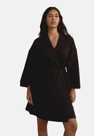 Femme aux longs cheveux foncés portant une robe portefeuille noire texturée avec de larges manches et une ceinture nouée à la taille, debout devant un fond blanc.