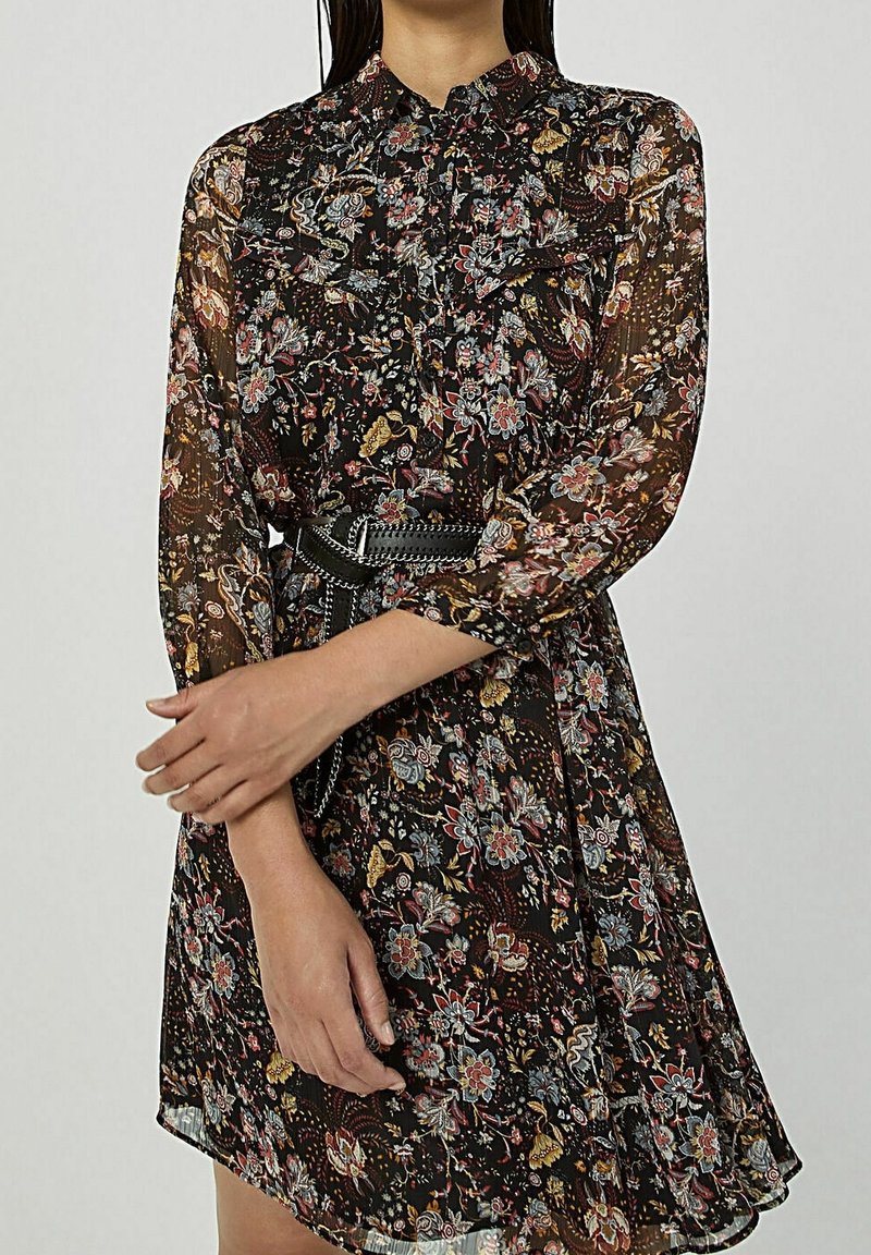 Robe noire à fleurs avec manches longues, col à volants, et taille cintrée accentuée par une ceinture texturée. Présente des motifs floraux colorés.