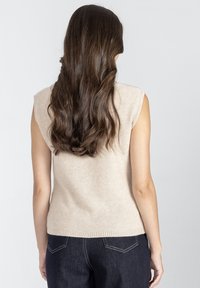 Beżowy bezrękawnik z dzianiny, okrągłym dekoltem, żeberkowym wykończeniem i miękką teksturą. Noszony z ciemnoniebieskimi spodniami dżinsowymi. Prosty fason, bez wzorów.