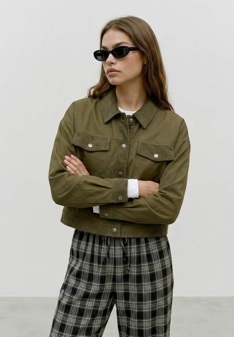 Femme aux longs cheveux bruns portant des lunettes de soleil noires, une veste boutonnée vert olive et un pantalon à carreaux noir et blanc, bras croisés.