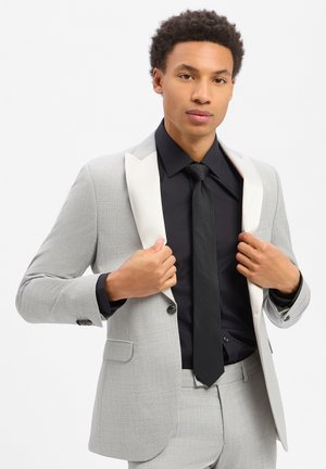 Giovane uomo che si sistema la giacca di un abito grigio chiaro con rever bianchi, indossando camicia nera e cravatta su sfondo semplice.