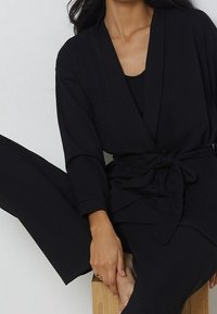 Completo loungewear nero con texture a coste, cardigan avvolgente con cintura e pantaloni tapered abbinati. Design semplice e moderno.