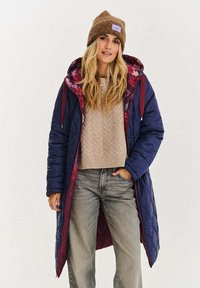 Pikowany granatowy długi płaszcz z bordowymi akcentami i kwiatową podszewką. W połączeniu z beżowym swetrem w warkocze i szarymi dżinsami. Na głowie brązowa czapka beanie.