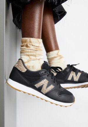 Nogi w czarnych tenisówkach New Balance z panterkowymi akcentami i kremowymi skarpetkami tie-dye na białym tle.