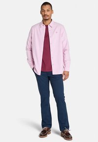 Ljusrosa skjorta med knappar över en vinröd T-shirt, matchad med mörkblå byxor och bruna skor med vita snören.