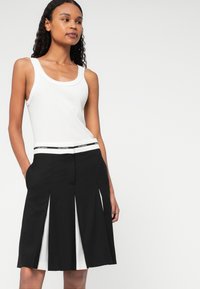 Kobieta w białej, bezrękawnikowej bluzce i czarnej spódnicy plisowanej z białymi panelami oraz markowym paskiem, stojąca na jednolitym tle.