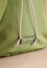 Zelená taška s hladkým povrchem, s růžovými stahovacími šňůrkami a obdélníkovou textilní etiketou s nápisem "Cath Kidston" ve zlatém písmu.