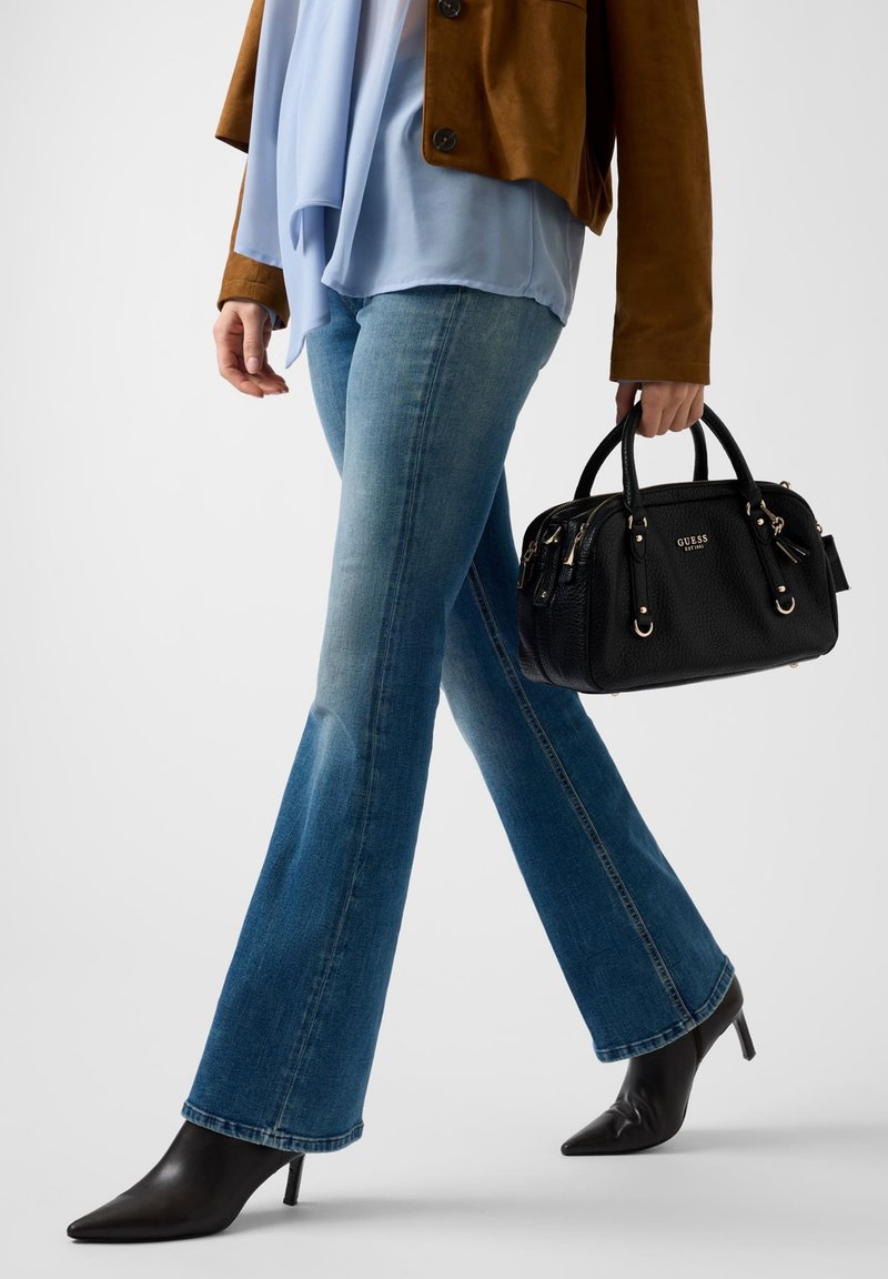 Donna che cammina indossando jeans svasati blu, stivali neri con tacco a punta, giacca marrone, camicetta azzurro chiaro, con una borsa nera Guess in mano.