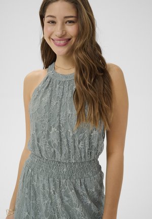 Mladá žena sa usmieva, má na sebe bezrukávové, svetlozelené čipkované šaty s vysokým golierom, s riaseným pásom a dlhými vlnitými vlasmi prehodenými cez jedno rameno.