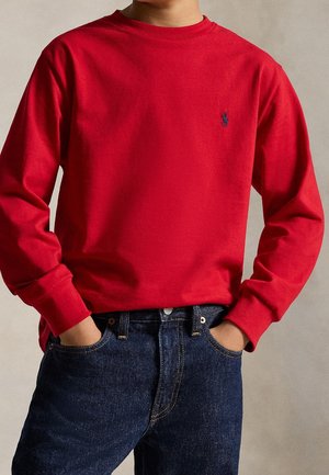Person iført en rød langærmet skjorte med et lille mørkt broderet logo på brystet, hænderne i lommerne på mørkeblå jeans.