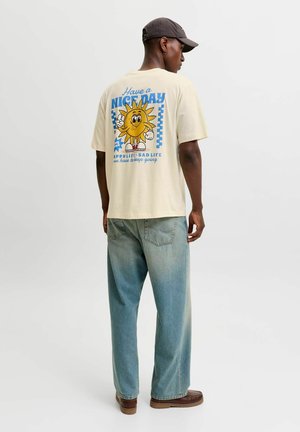 Homme portant un t-shirt beige avec un graphique coloré de soleil souriant et un texte au dos, jean bleu clair, chaussures marron et casquette foncée, debout de côté.