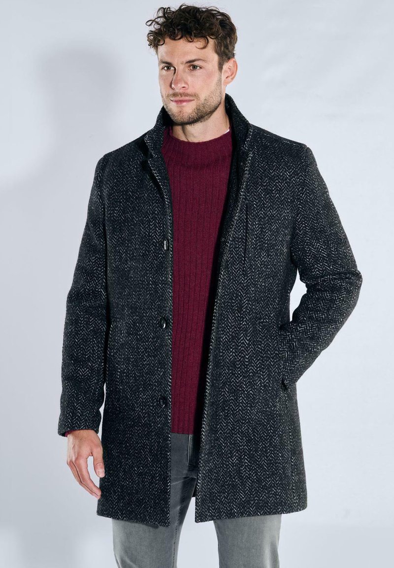 Mantel aus Wollmischung in Schwarz mit Fischgrätmuster, ausgestattet mit einem Reverskragen und Knopfverschluss, kombiniert mit einem bordeauxroten, gerippten Pullover.