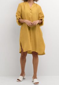 Frau, gekleidet in einem lockeren, senfgelben knielangen Kleid mit hochgerollten Ärmeln und weißen, offenen Blockabsatz-Mules vor einem einfarbigen Hintergrund.