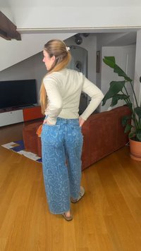 Mujer con el pelo largo sujetado con una pinza, lleva un suéter beige y vaqueros de pierna ancha con estampado azul, de pie con las manos en los bolsillos traseros en una sala de estar.